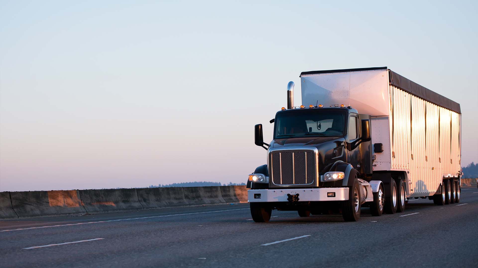Black semi truck with enclosed trailer traveling on a divided highway at sunset